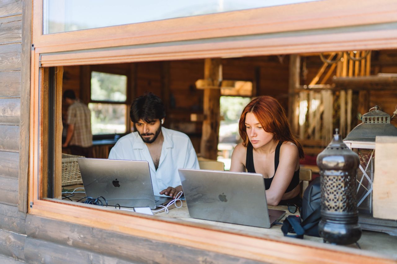 A man and woman working on laptops inside a rustic wooden cabin, sharing ideas.